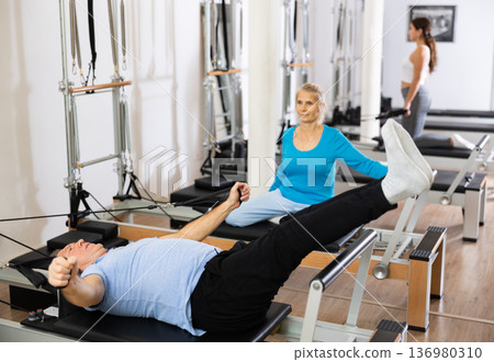 Focused mature male pensioner doing Pilates exercise for back on reformer bed equipment in rehabilitation center Focused mature male pensioner doing Pilates exercise for back on reformer bed equipment in rehabilitation center 136980310