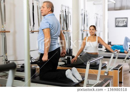 Focused mature male doing Pilates exercises for triceps while pulling bands on reformer bed at gym Focused mature male doing Pilates exercises for triceps while pulling bands on reformer bed at gym 136980318