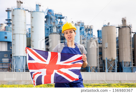 Portrait of upset girl in construction overalls with the Great Britain flag on the background of modern factory Portrait of upset girl in construction overalls with the Great Britain flag on the background of modern factory 136980367