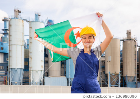Happy girl in work clothes and hardhat with flag of algeria standing in front of industrial scenery Happy girl in work clothes and hardhat with flag of algeria standing in front of industrial scenery 136980390