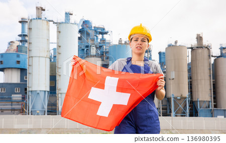 Worried girl in work clothes and hardhat with flag of switzerland standing in front of industrial scenery 136980395