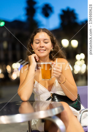 On summer outside of cafe, girl is relaxing at table, drinking chilled beer. On summer outside of cafe, girl is relaxing at table, drinking chilled beer. 136980628