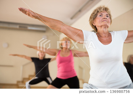 Group of aged women standing in warrior II pose in fitness room Group of aged women standing in warrior II pose in fitness room 136980756