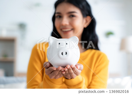 Young Asian woman holding piggy bank and sitting on couch in living room at home Young Asian woman holding piggy bank and sitting on couch in living room at home 136982016