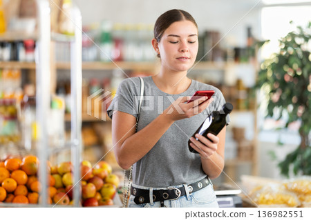 In shop youth girl scan QR code and take reading olive oil package bottle using phone camera 136982551