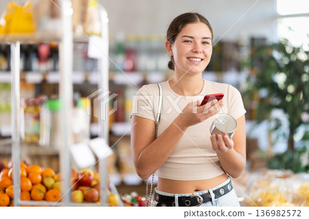 European girl customer at shop scans QR code on tin cans of preserves using phone camera European girl customer at shop scans QR code on tin cans of preserves using phone camera 136982572