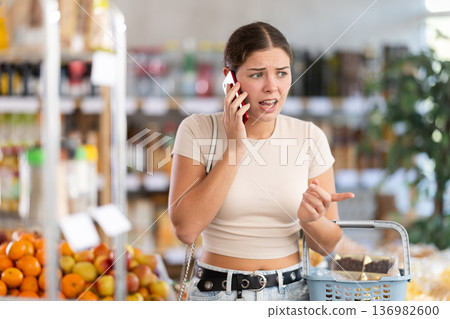 Angry young woman choosing groceries while talking on phone Angry young woman choosing groceries while talking on phone 136982600