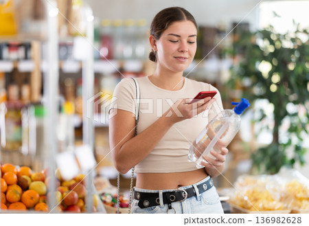 European young woman customer at shop scans QR code on soda bottle using phone camera 136982628