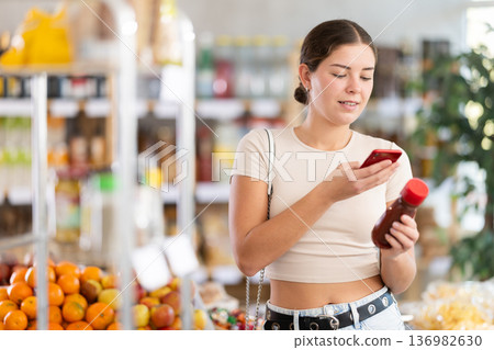 European young woman customer at shop scans QR code on ketchup bottle using phone camera 136982630