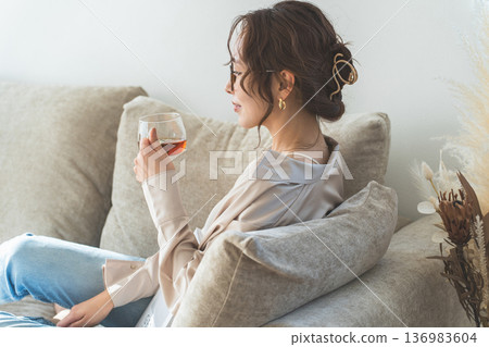 一位穿著時尚的亞洲女性坐在沙發上喝茶。 一位穿著時尚的亞洲女性坐在沙發上喝茶。 136983604