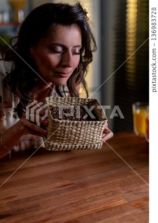 Woman presents detailed handcrafted woven container in kitchen setting, Woman gently lifts and displays meticulously woven artisan basket highlighting craftsmanship details Woman presents detailed handcrafted woven container in kitchen setting, Woman gently lifts and displays meticulously woven artisan basket highlighting craftsmanship details 136983728