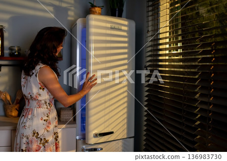 Woman seeks comfort in kitchen, Ambient scene of woman retrieving snack in dim light, Calm apartment setting with floral dressed woman exploring refrigerated items at night 136983730