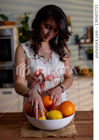 Selecting fruit in morning light, Soft light as woman selects citrus from arranged fruit bowl, Woman hand picks citrus fruit from bowl during gentle morning illumination Selecting fruit in morning light, Soft light as woman selects citrus from arranged fruit bowl, Woman hand picks citrus fruit from bowl during gentle morning illumination 136983733