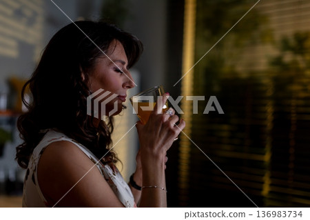 Woman savoring orange drink by window, freelance writer taking quiet latenight pause, reflective gaze, glass near lips, city lights outside, moody grain, intimate ambiance 136983734