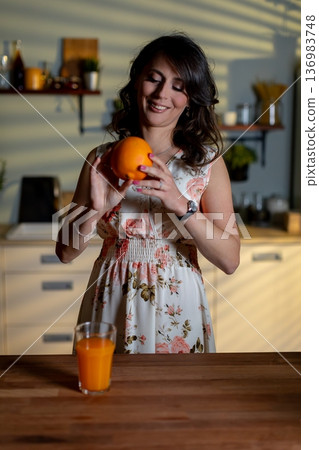 Joyful woman enjoying refreshment, Bright female with fruit and smile, Woman in floral dress with orange and juice, Happy lady with glass of juice and floral outfit in cozy kitchen Joyful woman enjoying refreshment, Bright female with fruit and smile, Woman in floral dress with orange and juice, Happy lady with glass of juice and floral outfit in cozy kitchen 136983748