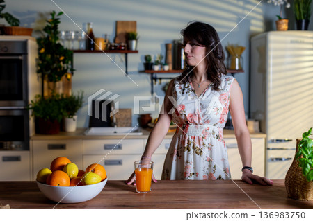 Serene scene of woman with fresh orange juice in warm kitchen environment, Calm moment captured as woman in floral dress savors healthy beverage by sunny kitchen window Serene scene of woman with fresh orange juice in warm kitchen environment, Calm moment captured as woman in floral dress savors healthy beverage by sunny kitchen window 136983750