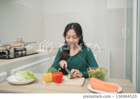 Young Woman Preparing Fresh Vegetables in Modern Kitchen, Slicing Cucumbers with Colorful Ingredients on Table, Healthy Cooking Concept Young Woman Preparing Fresh Vegetables in Modern Kitchen, Slicing Cucumbers with Colorful Ingredients on Table, Healthy Cooking Concept 136984520