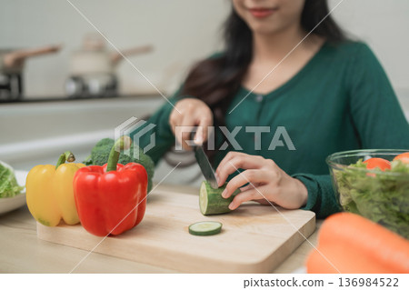 Woman Slicing Cucumber in Kitchen Surrounded by Fresh Vegetables and Ingredients for Healthy Meal Preparation and Cooking Woman Slicing Cucumber in Kitchen Surrounded by Fresh Vegetables and Ingredients for Healthy Meal Preparation and Cooking 136984522