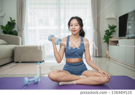 Joyful woman exercising at home with dumbbell while sitting on yoga mat in modern living room, showcasing a healthy lifestyle and fitness focus Joyful woman exercising at home with dumbbell while sitting on yoga mat in modern living room, showcasing a healthy lifestyle and fitness focus 136984528