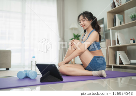 Young woman exercising at home with an iPad, dumbbells, and water bottle, engaging in a healthy lifestyle and fitness routine in a modern living room Young woman exercising at home with an iPad, dumbbells, and water bottle, engaging in a healthy lifestyle and fitness routine in a modern living room 136984548