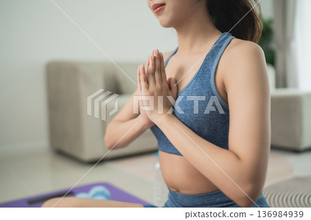 Young woman meditating indoors with folded hands while practicing yoga in sportswear, showcasing calmness and focus in a serene living space environment Young woman meditating indoors with folded hands while practicing yoga in sportswear, showcasing calmness and focus in a serene living space environment 136984939