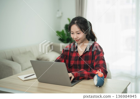 Young woman working on laptop in cozy living room, wearing casual plaid shirt, focused on tasks with plants and decorations in background Young woman working on laptop in cozy living room, wearing casual plaid shirt, focused on tasks with plants and decorations in background 136984945