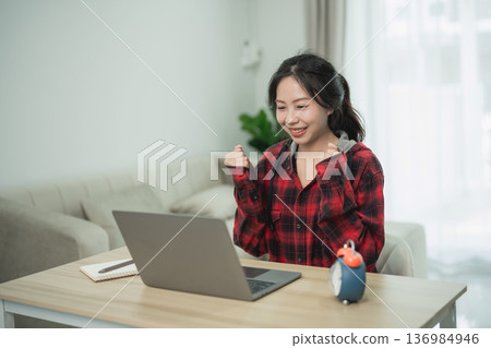 Young woman celebrating success at home while working on a laptop in casual attire, expressing joy and achievement in a cozy workspace Young woman celebrating success at home while working on a laptop in casual attire, expressing joy and achievement in a cozy workspace 136984946
