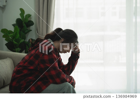 Young woman in casual outfit sitting on couch, holding her forehead in distress, reflecting on personal struggles in a modern and bright room setting Young woman in casual outfit sitting on couch, holding her forehead in distress, reflecting on personal struggles in a modern and bright room setting 136984969