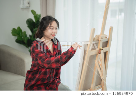 Young woman in casual attire painting on canvas indoors, surrounded by natural light, focusing on creativity and artistic expression in a cozy environment Young woman in casual attire painting on canvas indoors, surrounded by natural light, focusing on creativity and artistic expression in a cozy environment 136984970