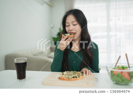 Young woman enjoying delicious pizza in a cozy dining room with salad and beverage on the table, creating a warm and inviting atmosphere Young woman enjoying delicious pizza in a cozy dining room with salad and beverage on the table, creating a warm and inviting atmosphere 136984974
