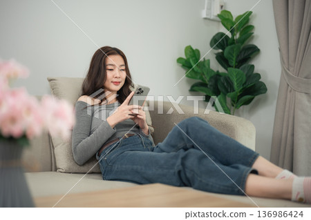 Relaxed young woman using smartphone seated on a cozy couch in a stylish living room with green plants and soft-colored decor for lifestyle imagery Relaxed young woman using smartphone seated on a cozy couch in a stylish living room with green plants and soft-colored decor for lifestyle imagery 136986424