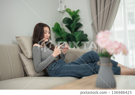 Young woman relaxing on couch while using smartphone in cozy living room with indoor plants and soft natural sunlight filtering through curtains Young woman relaxing on couch while using smartphone in cozy living room with indoor plants and soft natural sunlight filtering through curtains 136986425
