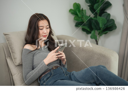 Young woman using smartphone while relaxing on couch in modern living room with indoor plants and cozy atmosphere for lifestyle or technology concept 136986426