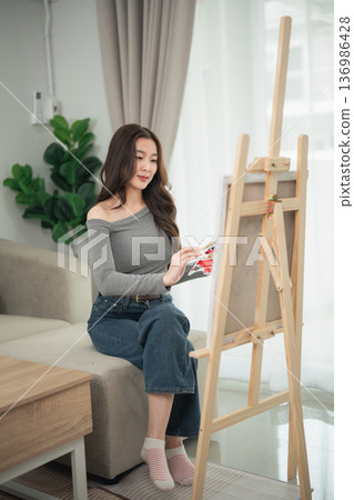 Young woman painting on canvas in modern living room with natural light, casual outfit, artistic expression, creative process captured in cozy environment Young woman painting on canvas in modern living room with natural light, casual outfit, artistic expression, creative process captured in cozy environment 136986428