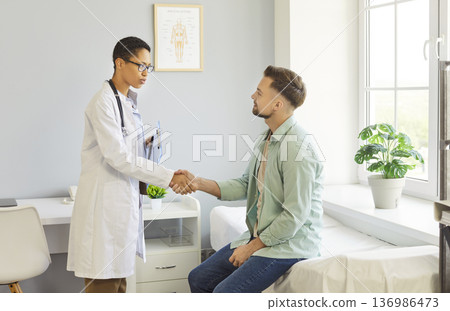 Clinic room meeting, female doctor shaking hands with male patient sitting on examination bed Clinic room meeting, female doctor shaking hands with male patient sitting on examination bed 136986473