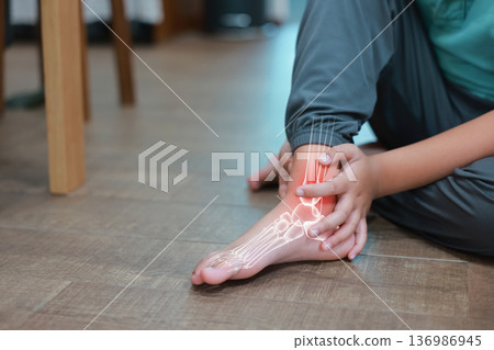 Little boy suffering from ankle joint pain with red x-ray bone graphic overlay, child touching foot feeling ache from injury, sprain, fracture or accidental fall while playing at home. 136986945