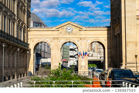 Scenic sunny view of the historic stone archway entrance with a clock and French flags at the Gare du Nord train station in Paris, Ile de France, France. Scenic sunny view of the historic stone archway entrance with a clock and French flags at the Gare du Nord train station in Paris, Ile de France, France. 136987593
