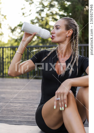 Athletic woman drinking water from sports bottle after outdoor workout outside in park. Fit female in black activewear resting in park, healthy lifestyle, hydration, fitness recovery Athletic woman drinking water from sports bottle after outdoor workout outside in park. Fit female in black activewear resting in park, healthy lifestyle, hydration, fitness recovery 136987946