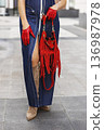 Close up of fashionable woman wearing denim maxi dress and red leather gloves holding stylish fringe handbag outdoors. Modern female street style with beige suede boots and bold color accessories 136987978