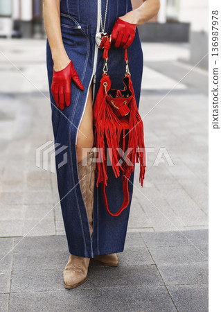 Close up of fashionable woman wearing denim maxi dress and red leather gloves holding stylish fringe handbag outdoors. Modern female street style with beige suede boots and bold color accessories 136987978