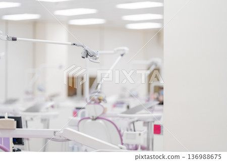The side view of a modern white dental light lamp and reflector equipment above a medical chair on a blurry background of a dental clinic. The side view of a modern white dental light lamp and reflector equipment above a medical chair on a blurry background of a dental clinic. 136988675