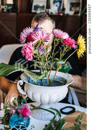 Man arranges bright aster flowers in ornate white pedestal urn. Sustainable mechanics, green living, garden-to-vase, environmentally conscious hobby. Man arranges bright aster flowers in ornate white pedestal urn. Sustainable mechanics, green living, garden-to-vase, environmentally conscious hobby. 136989487