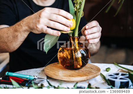 Hands arrange botanical elements in amber glass vase on wooden base. Men in floristry, modern masculine hobbies, artisan identity. Hands arrange botanical elements in amber glass vase on wooden base. Men in floristry, modern masculine hobbies, artisan identity. 136989489