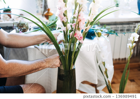 Man arranges gladiolus flowers in glass vase on white table. Men in floristry, breaking gender norms, male florist, creative masculinity, gender-neutral hobbies. 136989491