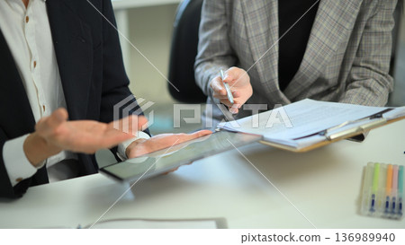 Close-up of coworkers discussing information using a tablet and documents at an office desk, showing data sharing, teamwork, and professional collaboration. Close-up of coworkers discussing information using a tablet and documents at an office desk, showing data sharing, teamwork, and professional collaboration. 136989940