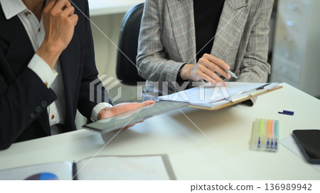 Close-up of coworkers discussing information using a tablet and documents at an office desk, showing data sharing, teamwork, and professional collaboration. 136989942