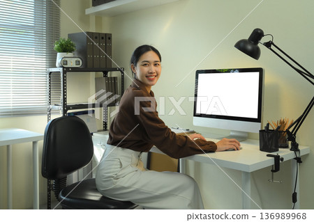 Office woman looking at camera and using blank screen computer monitor Office woman looking at camera and using blank screen computer monitor 136989968