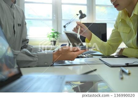 Coworkers exchanging information using a tablet and laptop during a business discussion, showing data sharing, teamwork, and professional Coworkers exchanging information using a tablet and laptop during a business discussion, showing data sharing, teamwork, and professional 136990020