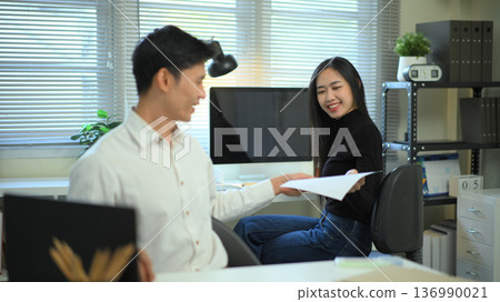 Colleagues exchange documents in a modern home office, discussing work progress with a computer and charts 136990021