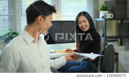 Colleagues exchange documents in a modern home office, discussing work progress with a computer and charts Colleagues exchange documents in a modern home office, discussing work progress with a computer and charts 136990073